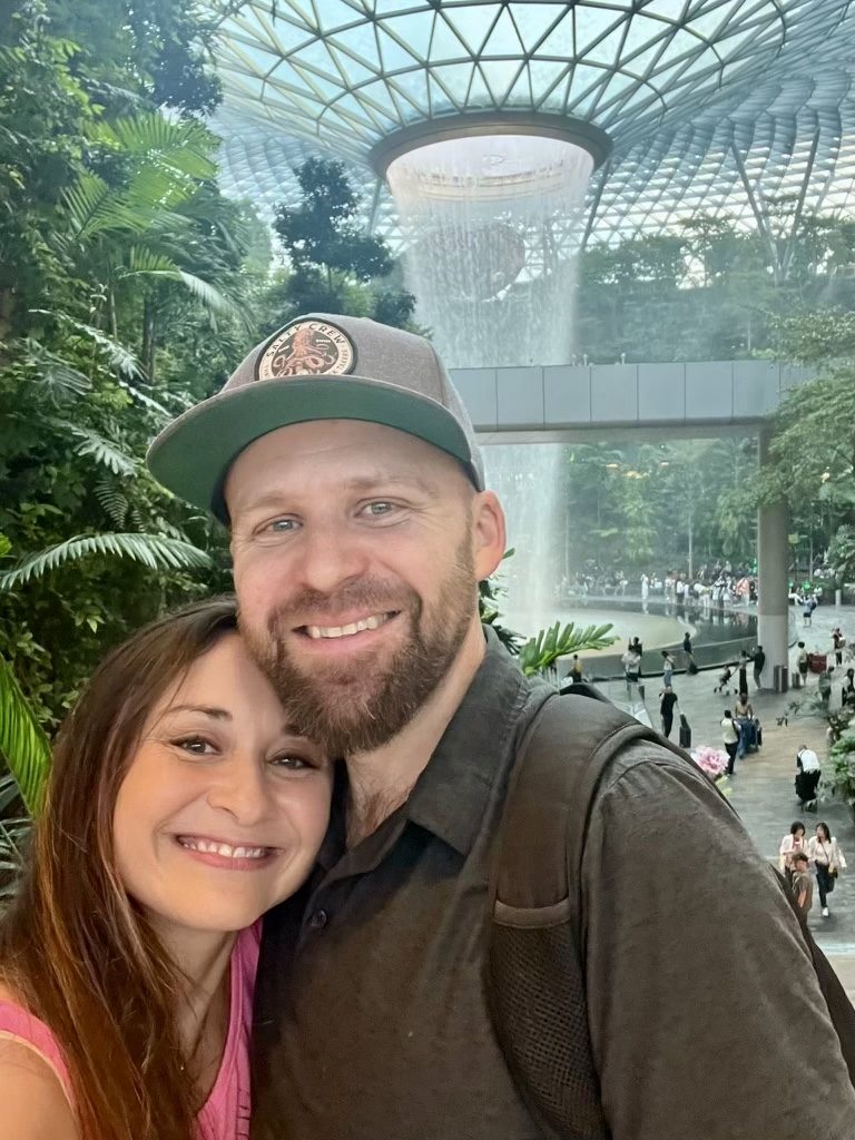 Jordan wearing a Salty Crew hat, smiling with a companion in front of the Jewel Changi Airport waterfall